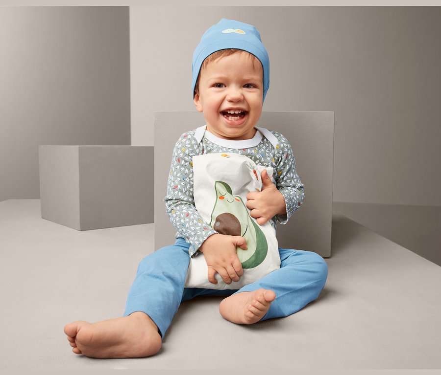 Lächelndes Kind sitzt und hält ein Rapssamen-Wärmekissen mit Avocado-Aufdruck.