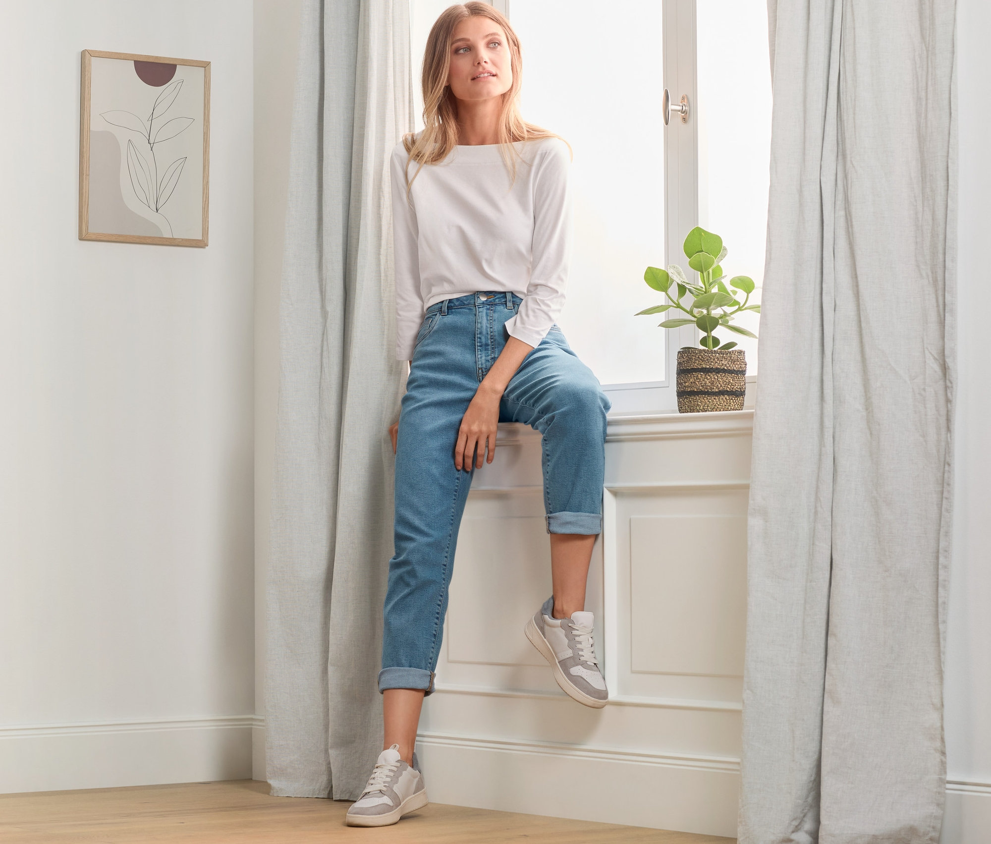 Blonde Frau sitzt im Fenster in Mom-Jeans und weißem Oberteil.