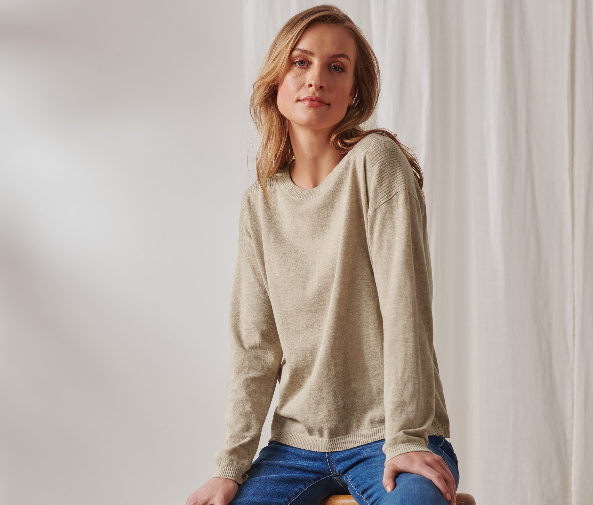 Blonde Frau sitzt auf einem Hocker und trägt einen beigen Feinstrickpullover mit Rippdetail und blaue Jeans.