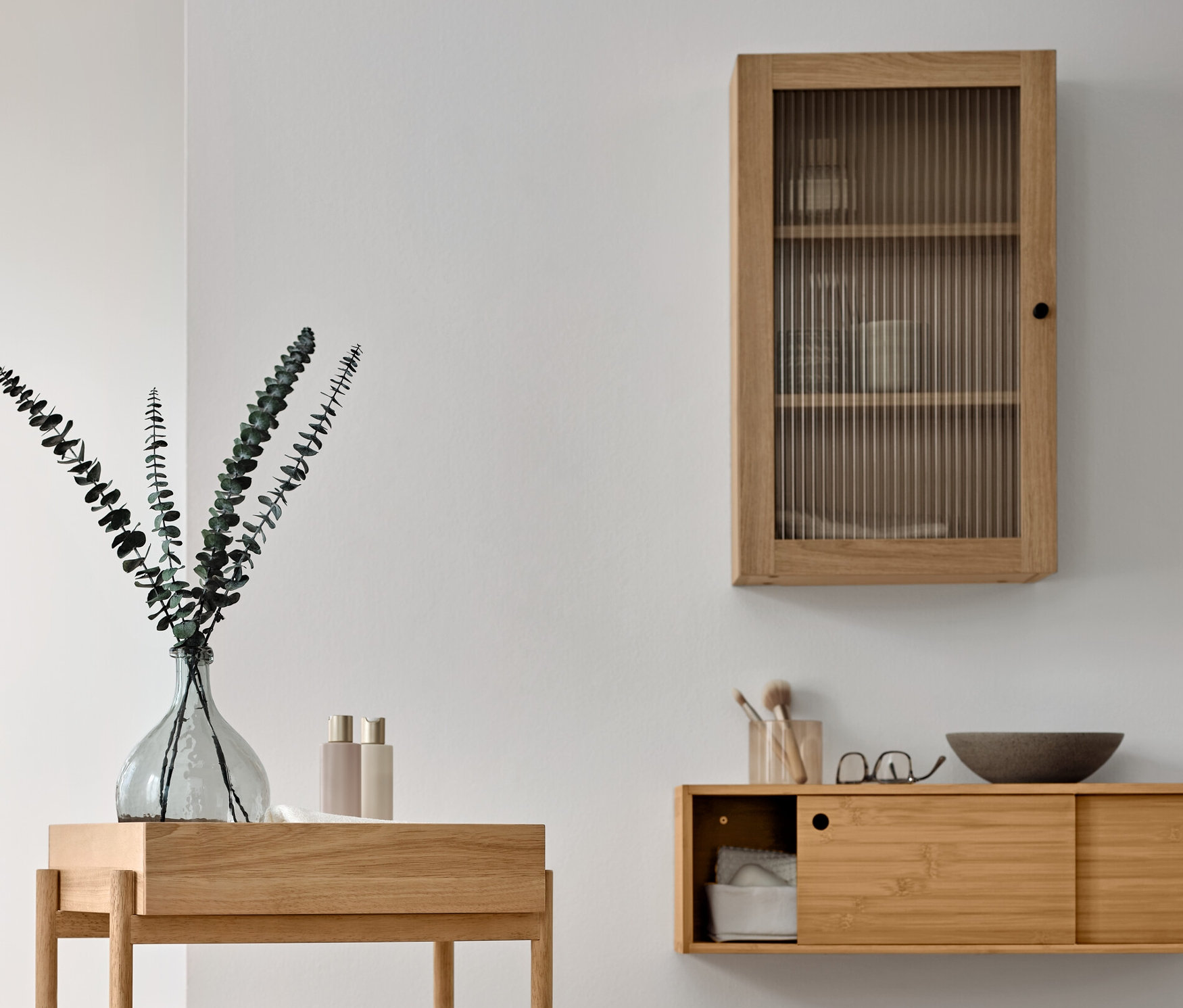 Einrichtung eines Badezimmers mit einem hölzernen Standregal mit 3 Fächern, einem Wandschrank mit Riffelglastür und einem Handtuchständer aus Massivholz.