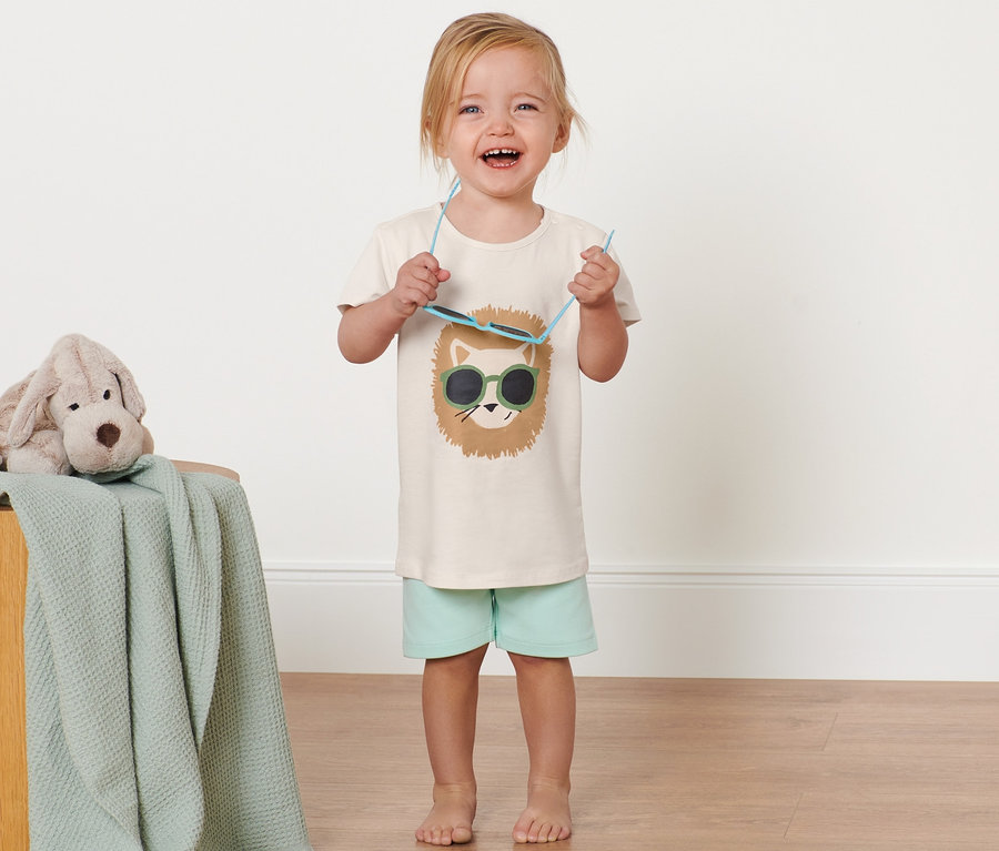 Ein lächelndes Kind hält eine Sonnenbrille und trägt ein Kinder-T-Shirt mit Wendepailletten. Ein Stoffhund liegt auf einem Holzschemel mit einer Decke darüber.