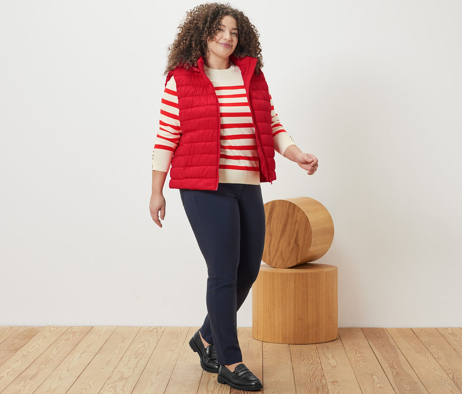 Frau geht, trägt einen rot gestreiften Feinstrickpullover, eine Steppweste mit Kapuze und eine dunkelblaue Bengalin-Hose.