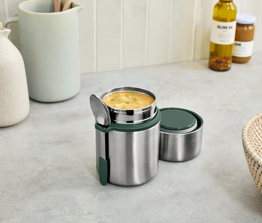 Ein Thermobehälter aus Edelstahl mit Suppe und einem Löffel, daneben ein Deckel. Im Hintergrund eine Kanne, ein Glas mit Olivenöl und ein Korb mit Obst.