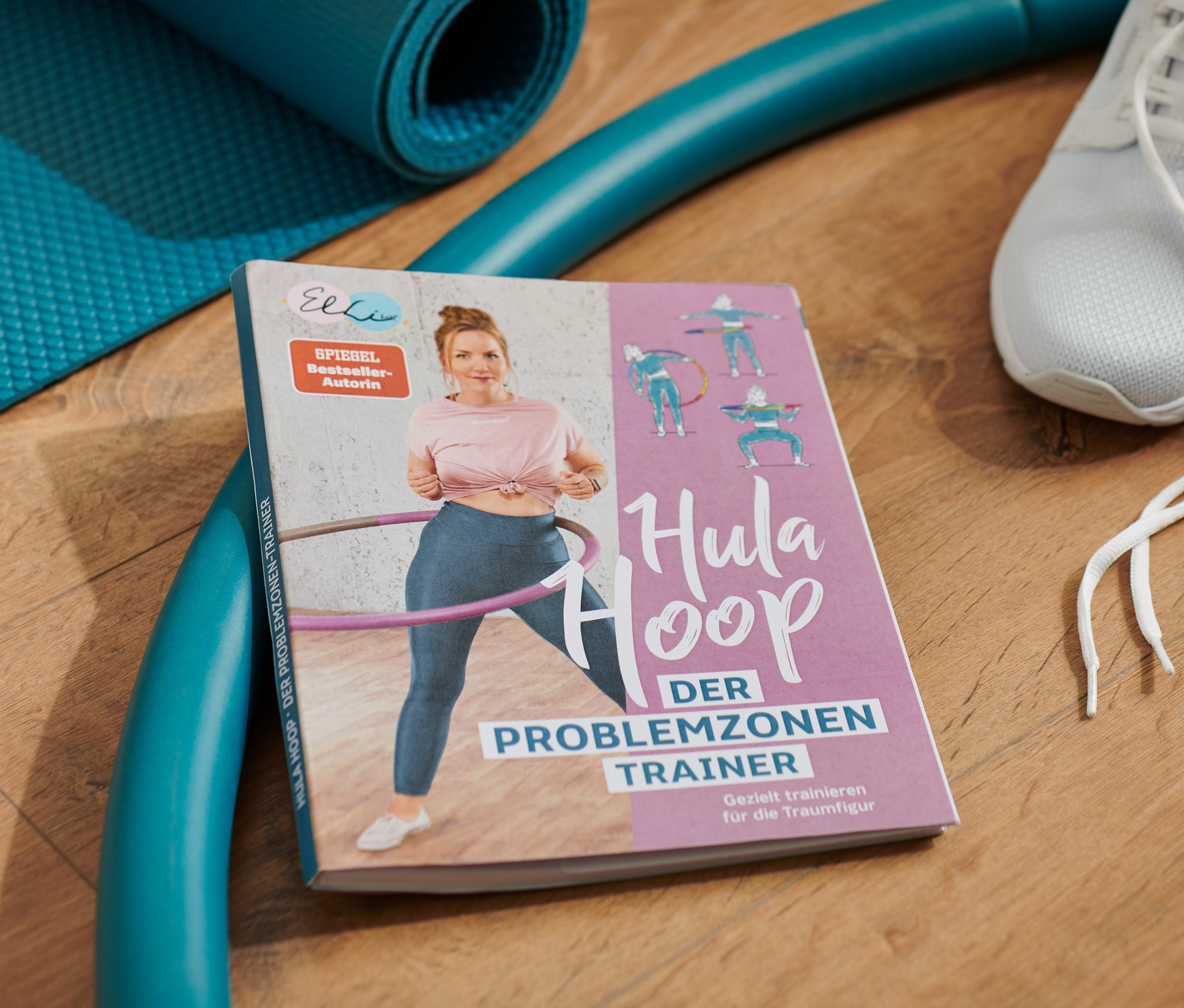 Auf einem Holzboden liegen ein Hula-Hoop-Buch, eine blaue Trainingsmatte, ein blauer Reifen und weiße Turnschuhe.