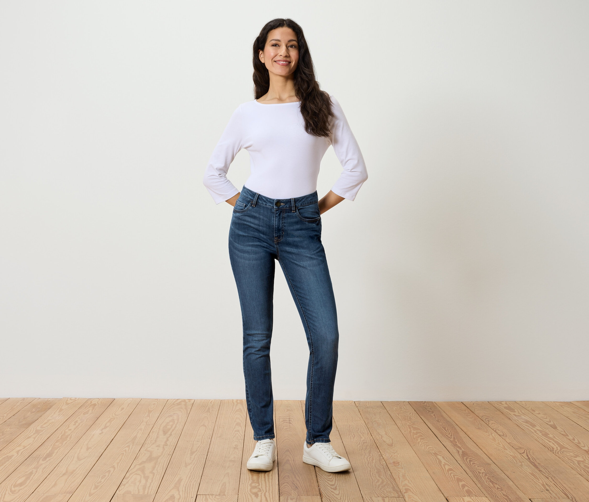 Frau steht mit den Händen in den Hüften und trägt blaue Jeans, eine weiße Bluse und weiße Turnschuhe.
