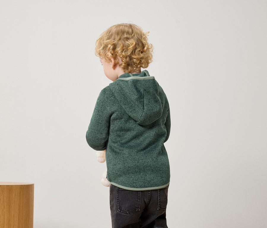 Ein Junge mit blonden Locken steht mit dem Rücken zur Kamera und trägt einen grünen Kapuzenpullover und schwarze Jeans.