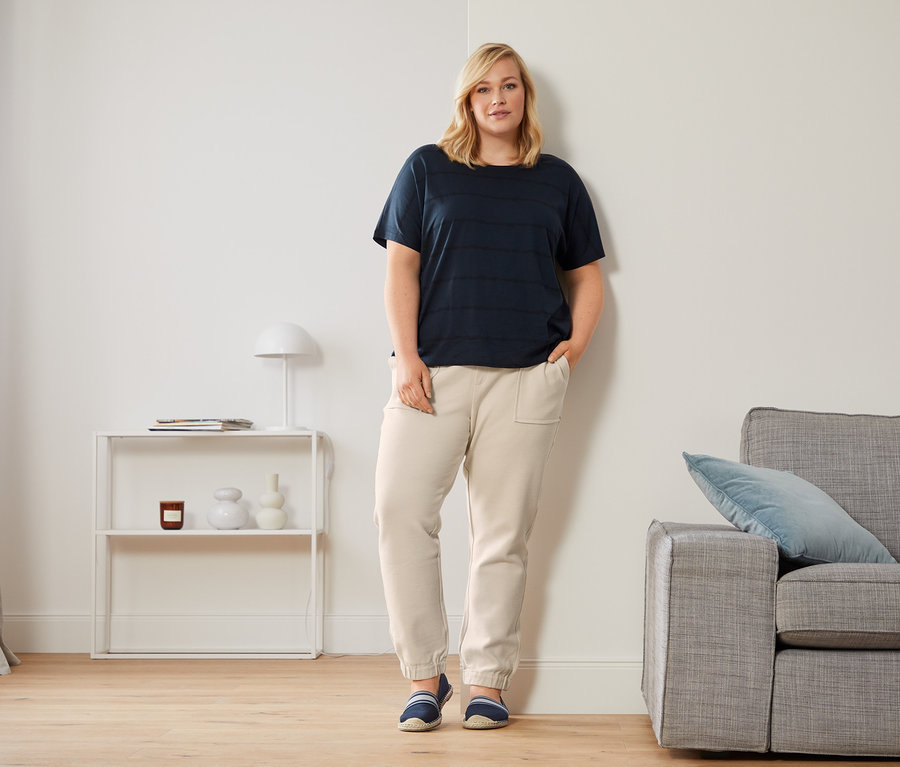 Frau lehnt an Wand, trägt dunkelblaues Shirt mit Rundhalsausschnitt, knöchellange Joggpants und Espadrilles.