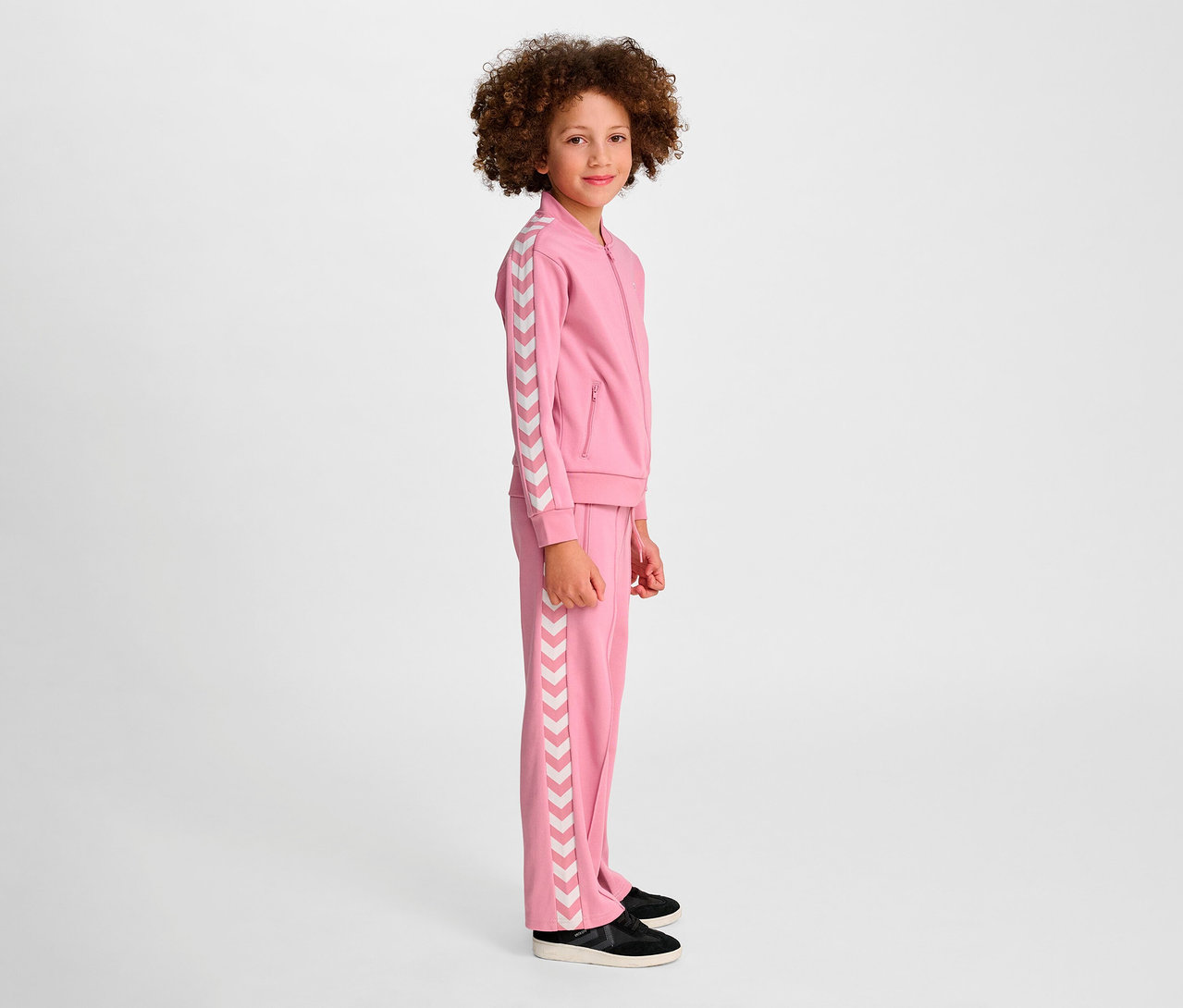 Mädchen steht seitlich und trägt einen rosa Hummel HMLJR Archive Reg Poly Zip Jacket Trainingsanzug.