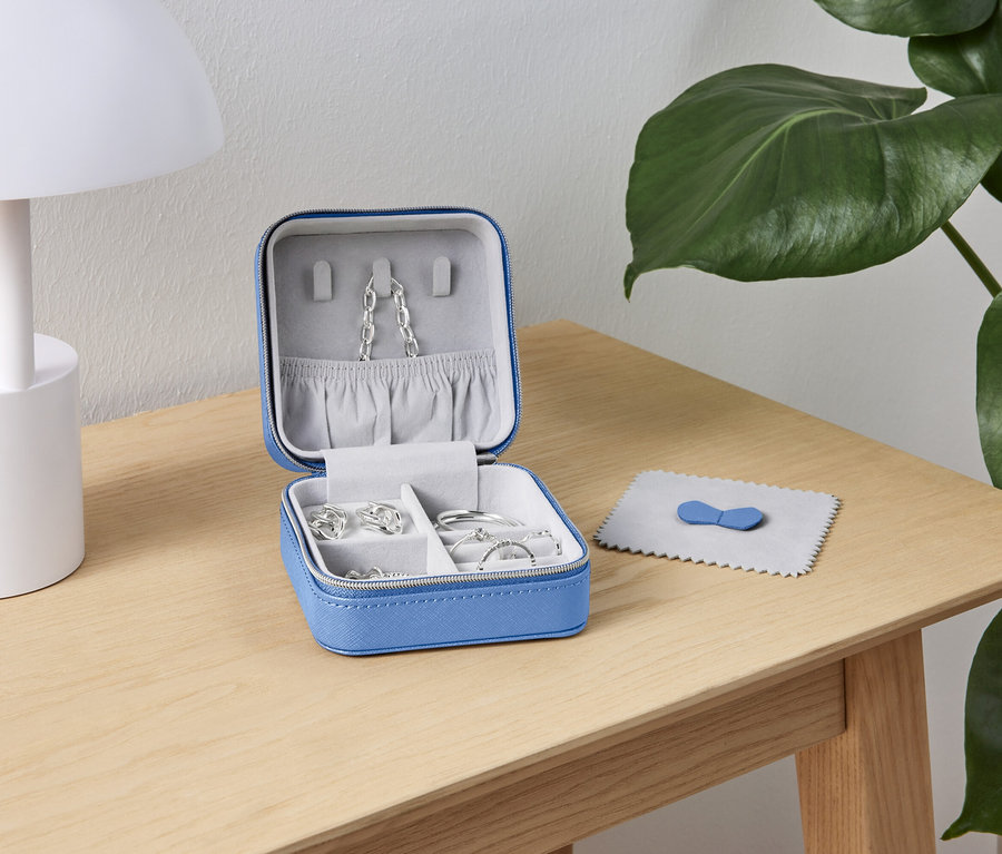 Ein offenes, blaues Schmuckkästchen mit silbernen Schmuckstücken steht auf einem Holztisch neben einer weißen Lampe und einer grünen Pflanze.