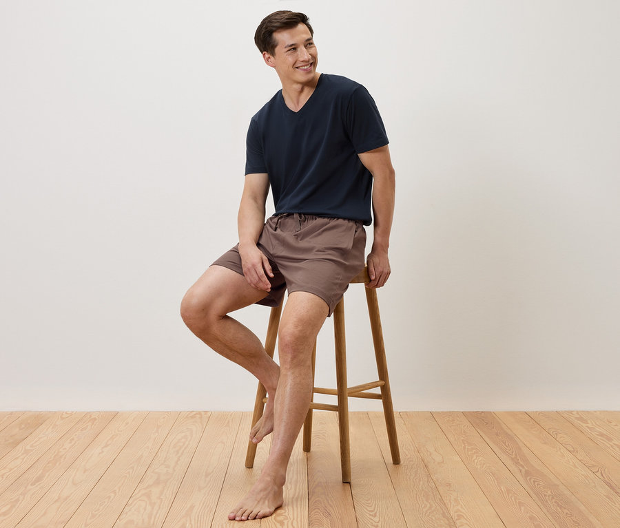 Mann sitzt barfuß auf einem Hocker, trägt ein marineblaues T-Shirt und braune Shorts.