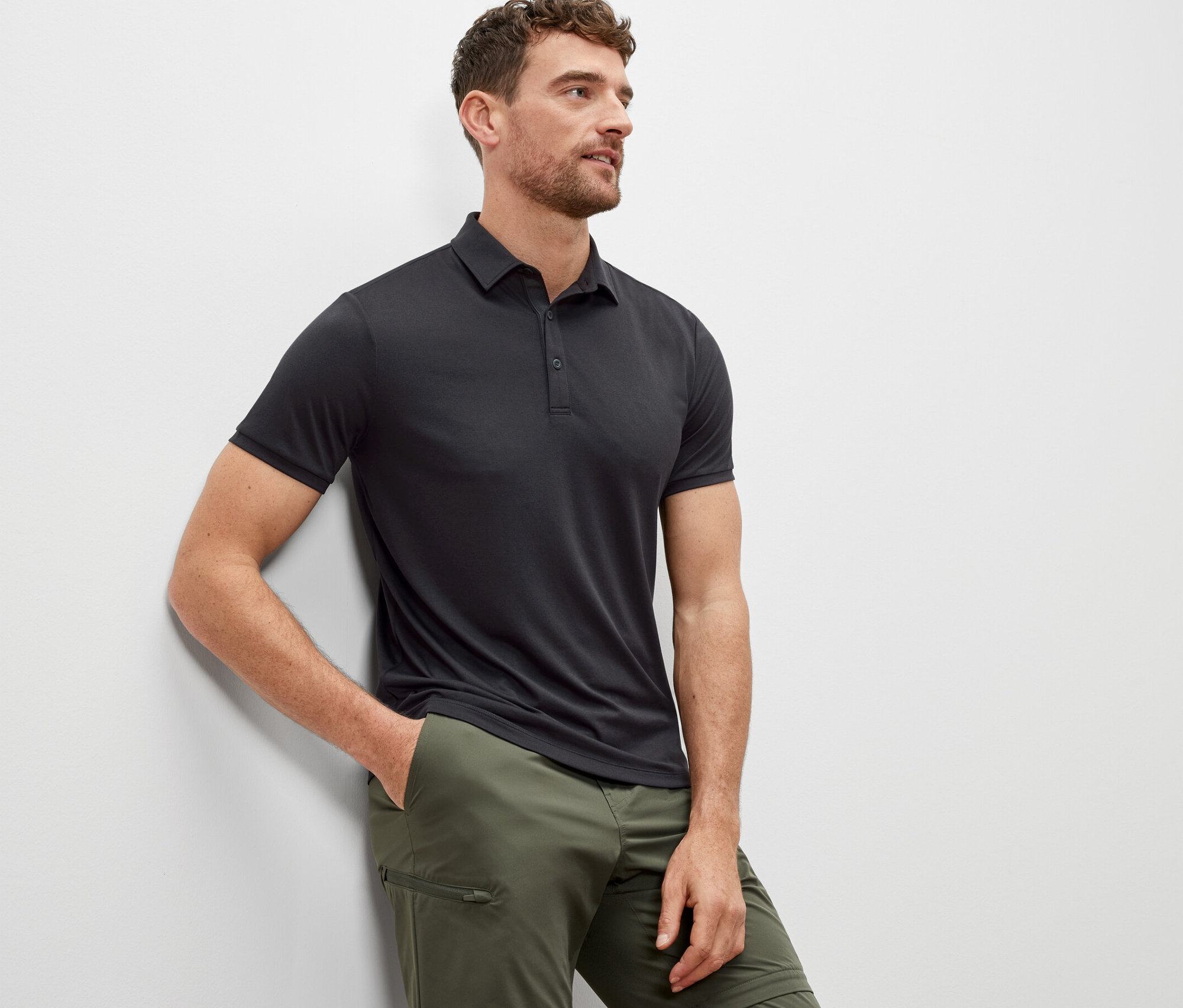 Mann lehnt an einer Wand und trägt ein anthrazitfarbenes Funktions-Poloshirt.