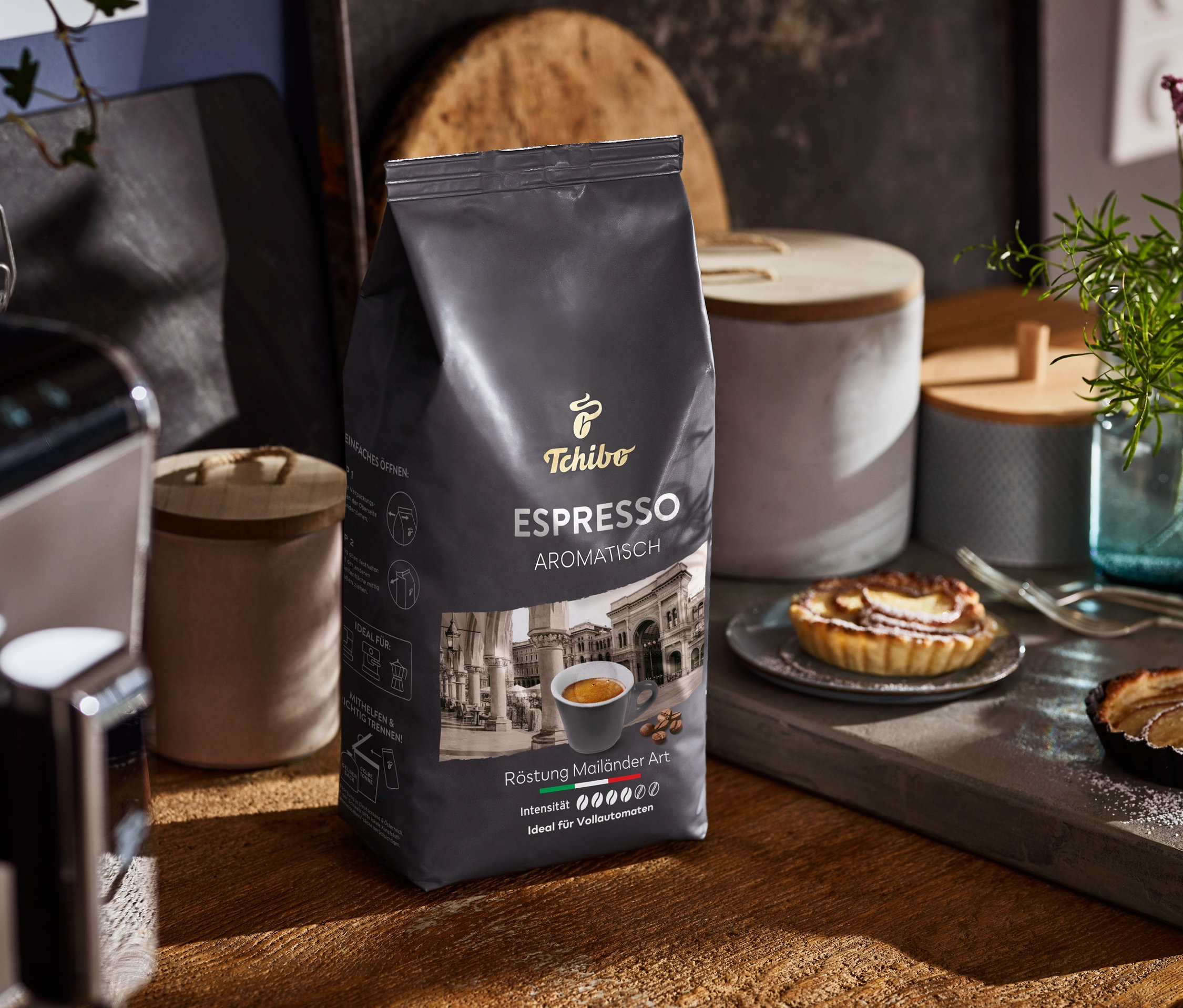 Eine Packung Espresso Aromatisch steht auf einem Holztisch neben einer Kaffeemaschine und Tellern mit Kuchen.