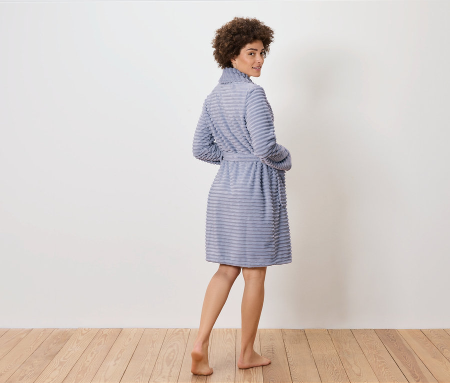 Frau blickt über die Schulter und posiert in einem blauen Bademantel auf einem Holzboden.