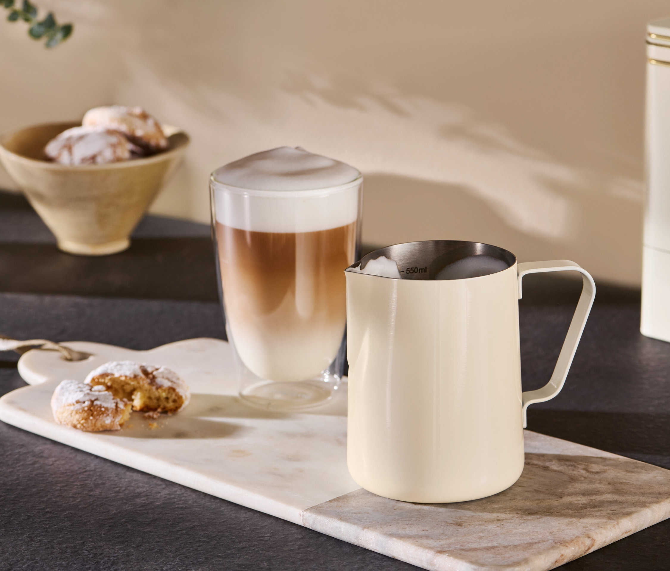 Ein beigefarbenes Milchkännchen steht neben einem Glas Kaffee auf einem Marmortablett.