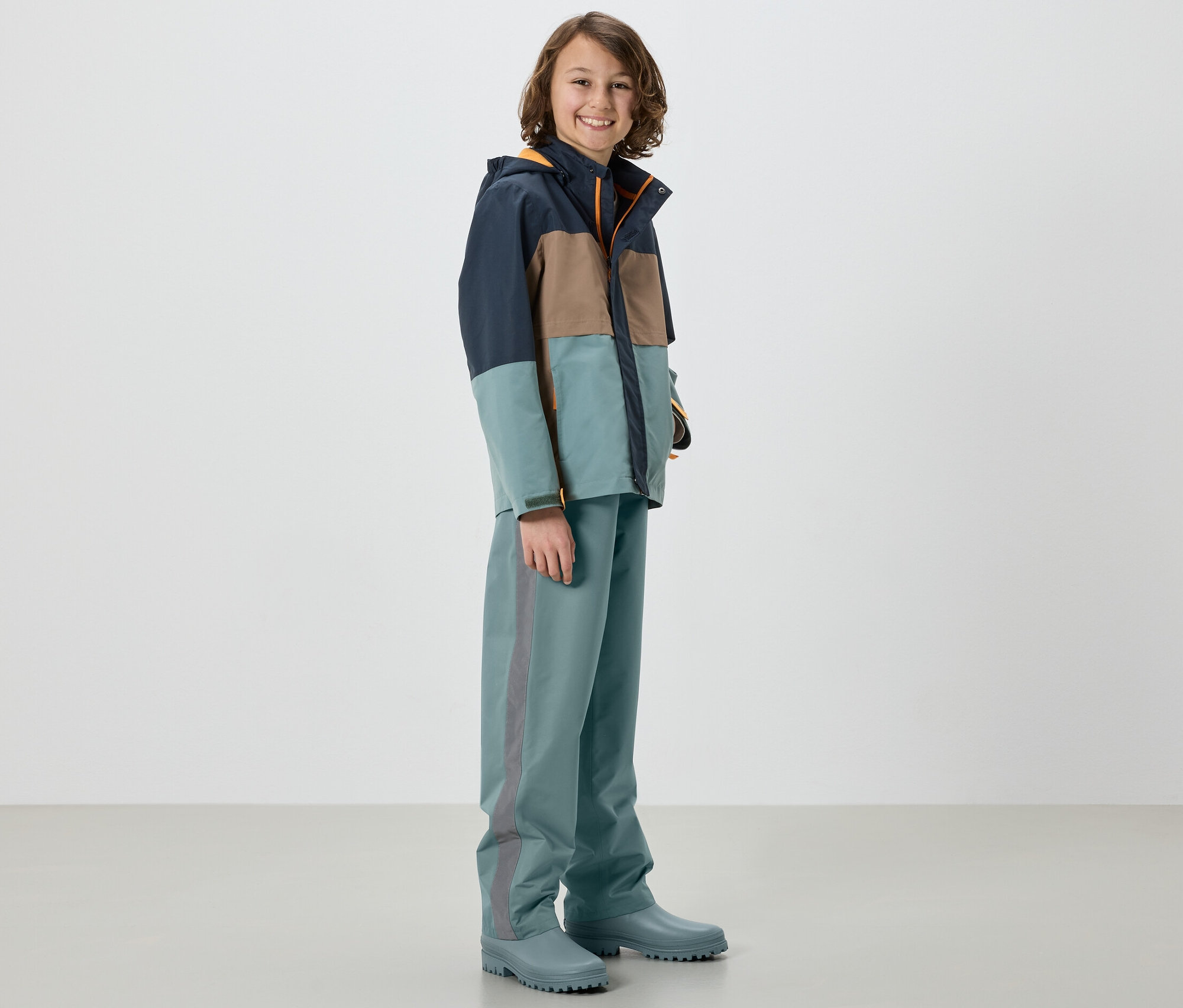 Ein Junge in einer Jacke und einer Kinder-Regenhose, Salbei, steht in Gummistiefeln.