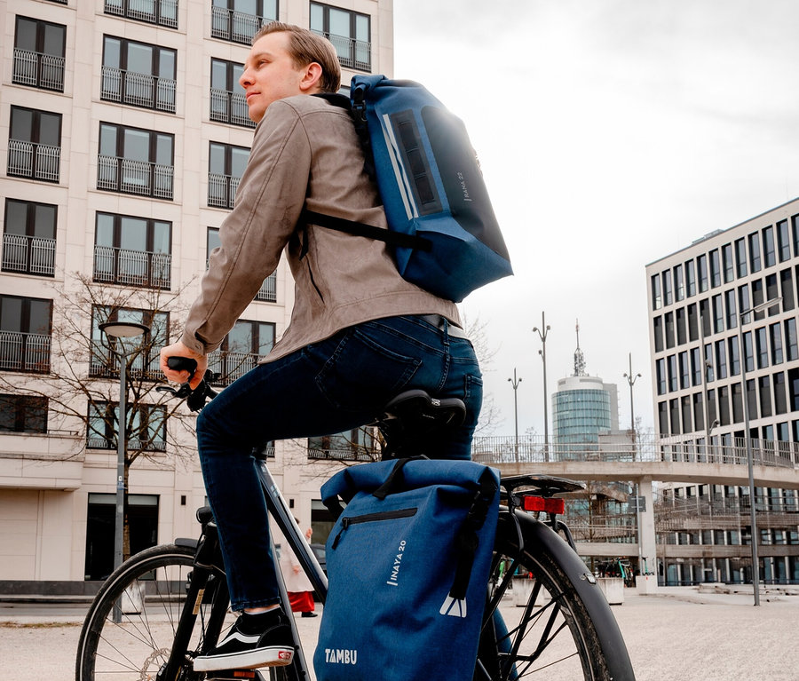 Mann fährt mit blauem Tambu Rolltop-Rucksack RANA auf dem Rücken Fahrrad.