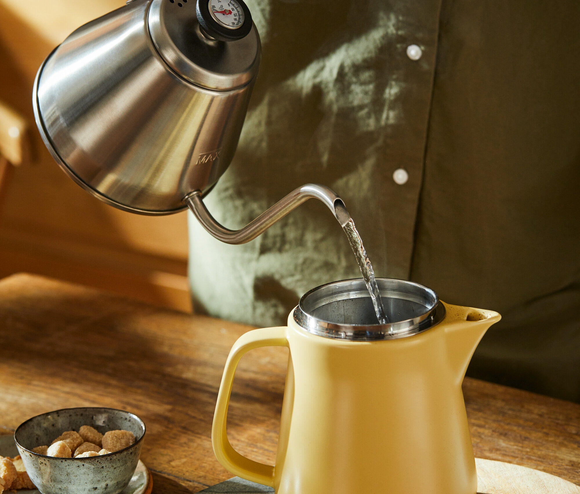 Person gießt Wasser aus einem Schwanenhalskessel in einen gelben Keramik-Kaffeebereiter.