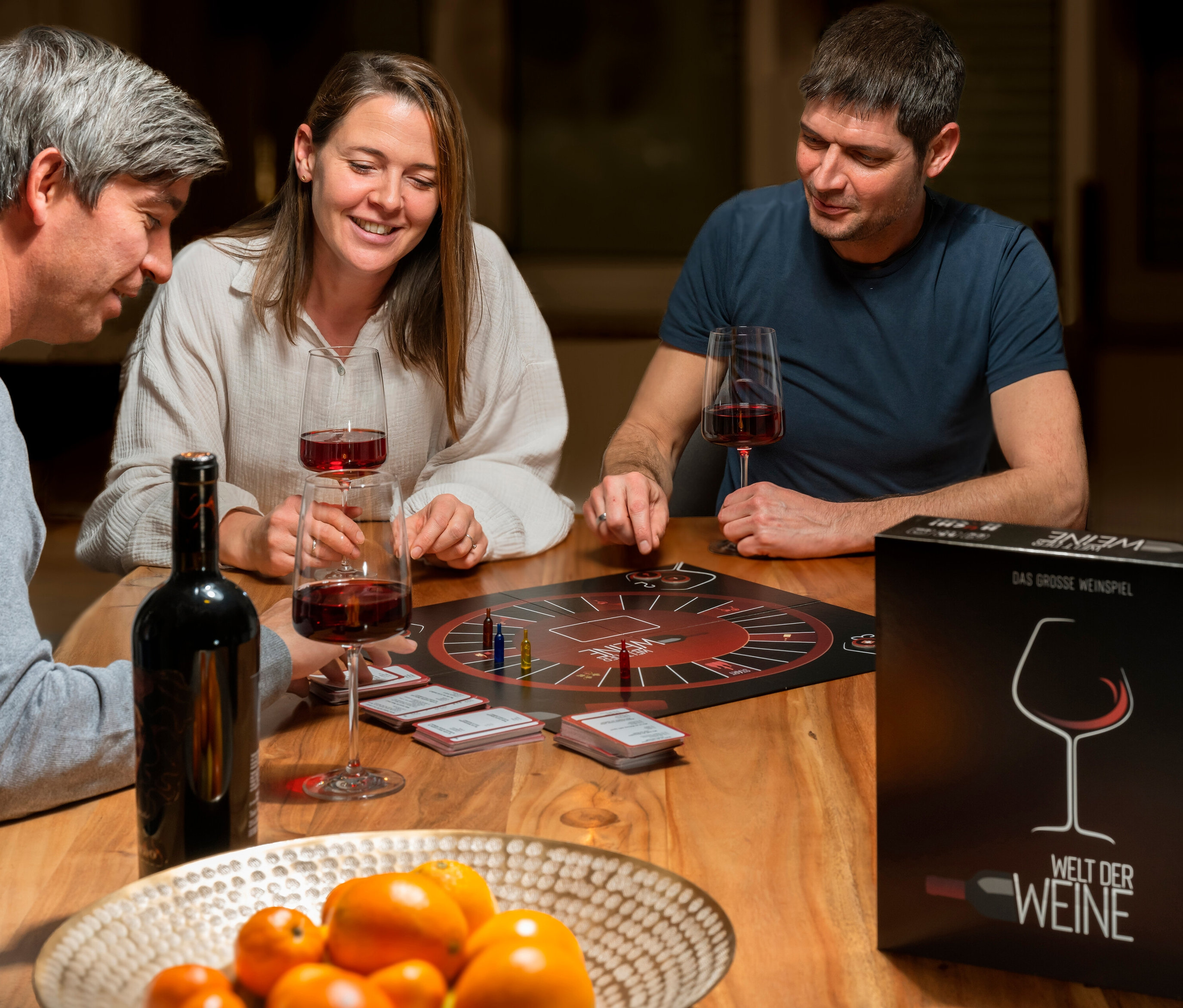 Drei Personen sitzen am Tisch und spielen das Spiel »Welt der Weine«.