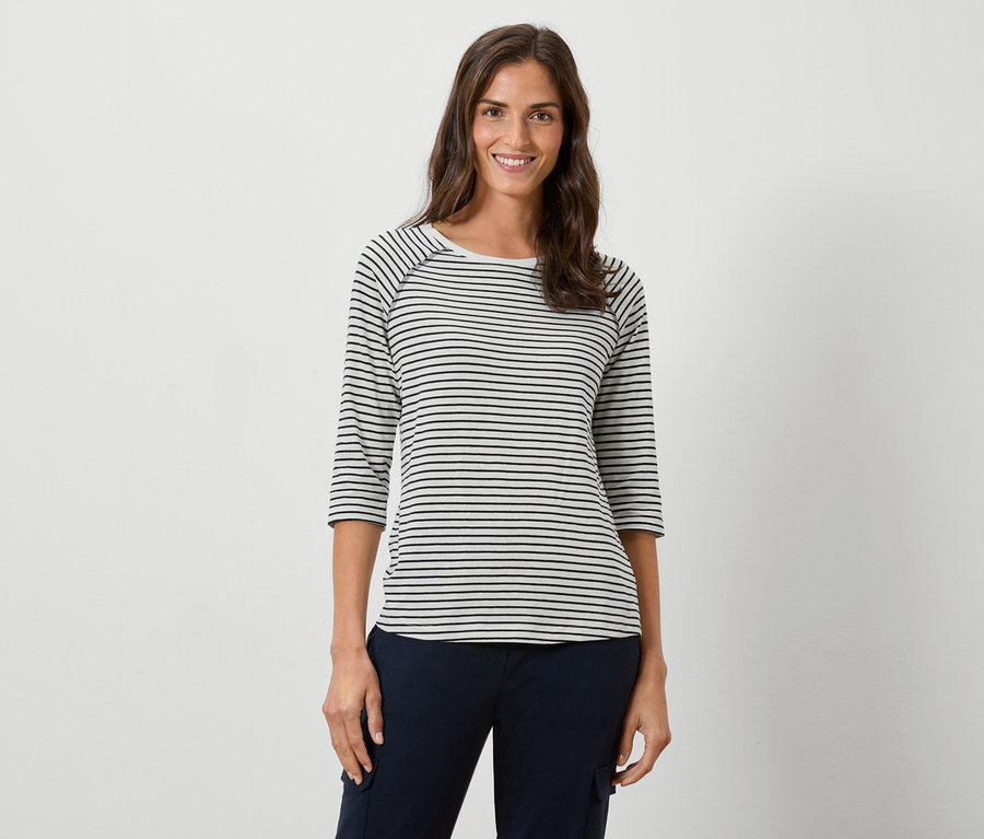 Frau trägt ein gestreiftes Langarmshirt und eine marineblaue Hose vor einem weißen Hintergrund.