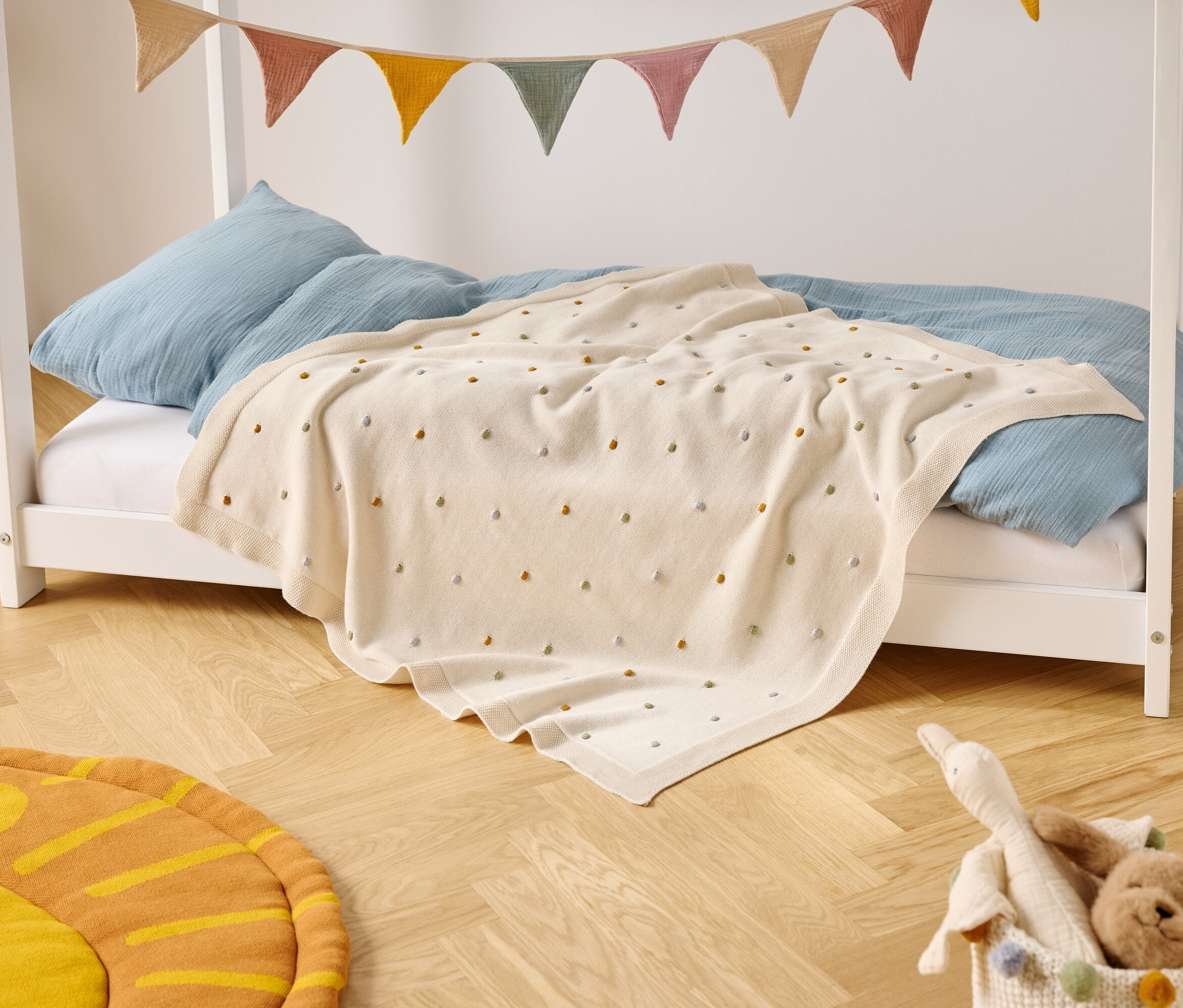 Ein Kinderbett mit blauen Kissen und einer cremefarbenen Decke mit farbigen Punkten steht auf einem Holzboden. Eine bunte Wimpelkette hängt über dem Bett.