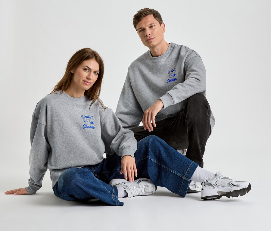 Eine Frau und ein Mann sitzen auf dem Boden und tragen graue Sweatshirts mit der blauen Aufschrift "Cheers".