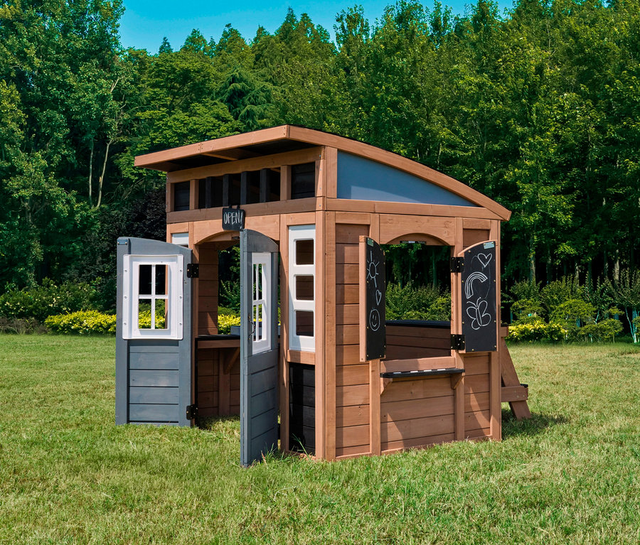 Das Spielhaus Trolli steht auf einer Wiese vor Bäumen. Es hat eine offene Tür und Fenster sowie eine Theke mit Kreidetafeln.