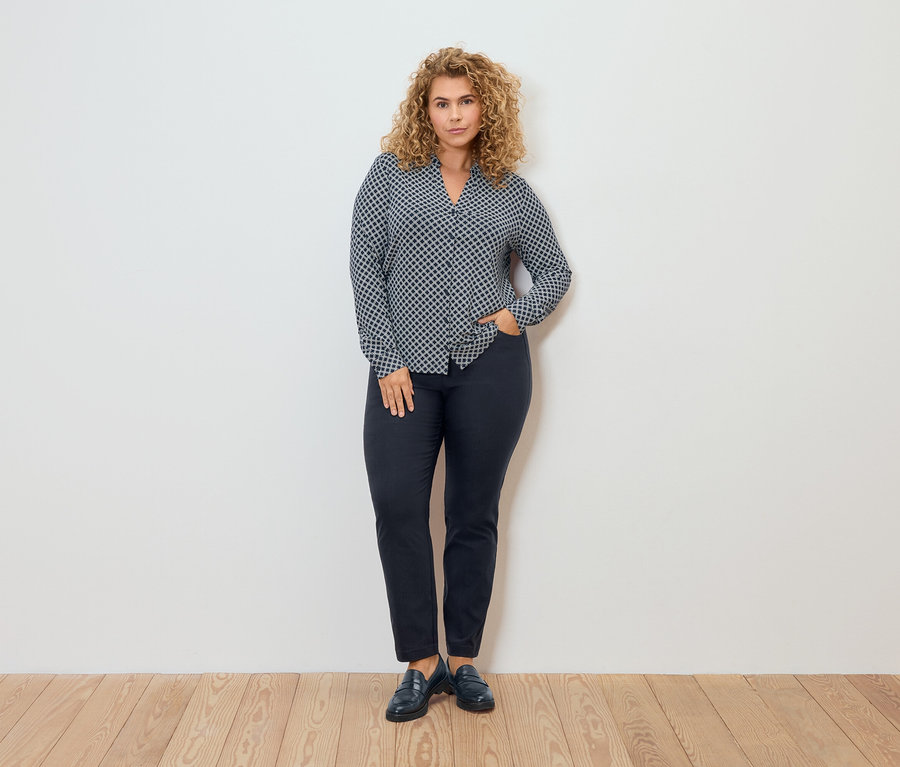 Frau in gemusterter Bluse und Bengalin-Hose posiert vor einer Wand.