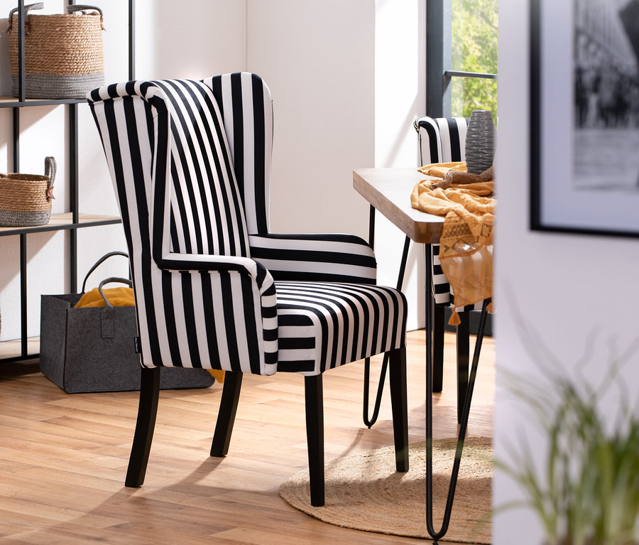 Ein schwarz-weiß gestreifter Hochlehner-Polsterstuhl »Hendrick« mit Armlehne steht neben einem Holztisch auf einem Teppich.