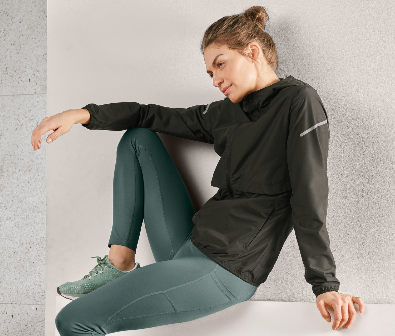 Eine Frau sitzt auf einer Kante und trägt eine schwarze Windprotection-Laufjacke, grüne Leggings und Turnschuhe.