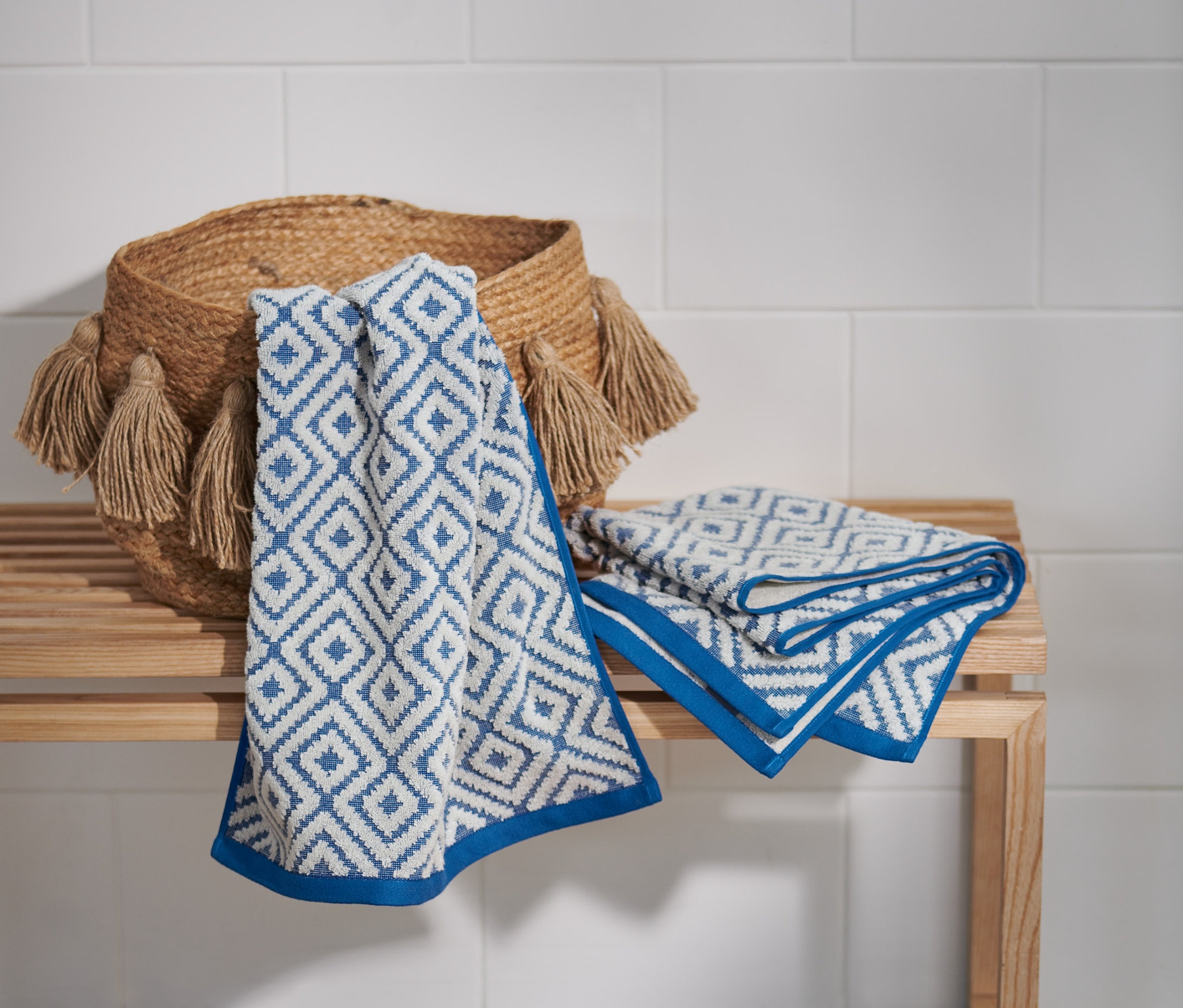 Zwei Jacquard-Handtücher mit blauem geometrischem Muster liegen auf einer Bank neben einem geflochtenen Korb mit Fransen.