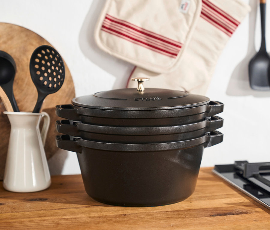 Staub Gusseisen-Kochtopf-Set auf einer Holzplatte neben einem Herd.