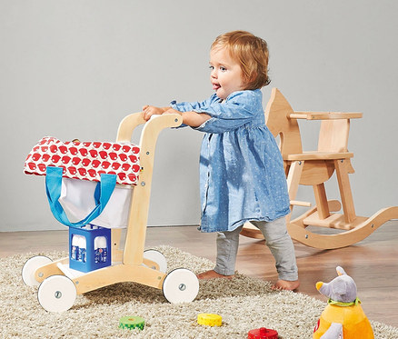 Kleinkind steht und schiebt einen weißen Pinolino Einkaufswagen »Erna«. Dahinter steht ein Schaukelpferd aus Holz.