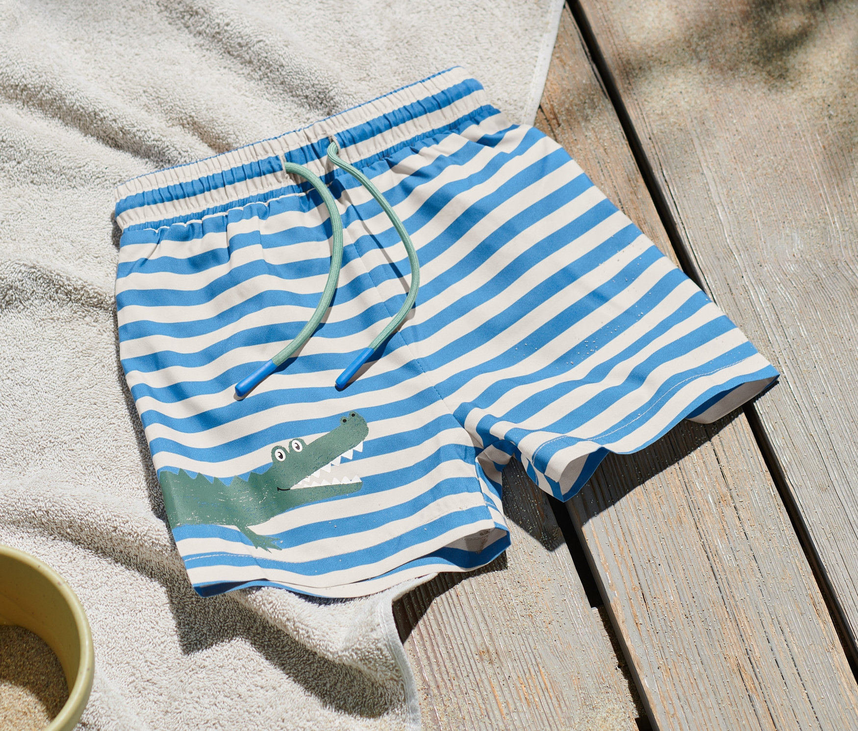 Kinder-Boardshorts mit blau-weißen Streifen und Krokodil-Motiv liegen auf einem Handtuch neben einem Sandspielzeug.