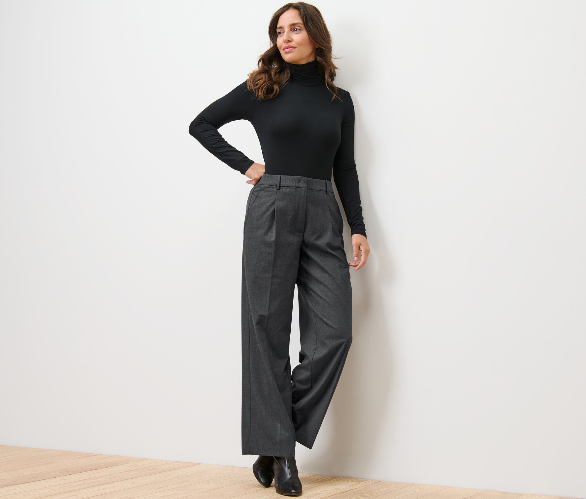 Frau posiert in schwarzem Rollkragenpullover und Komfort-Webhose an einer Wand.