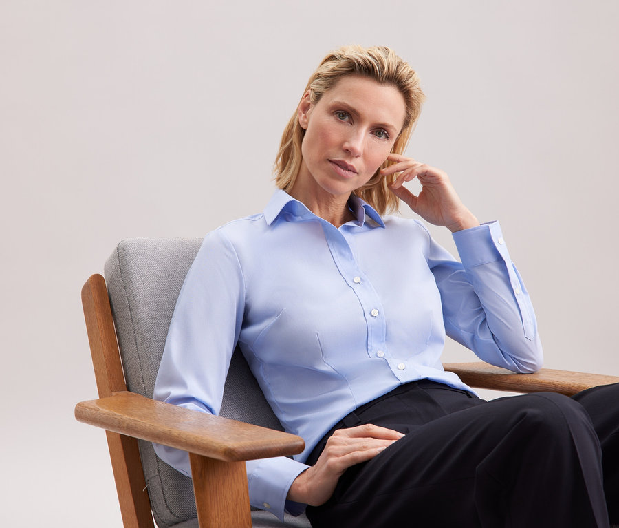Blonde Frau sitzt in einem grauen Sessel, trägt ein hellblaues Hemd und eine schwarze Hose.