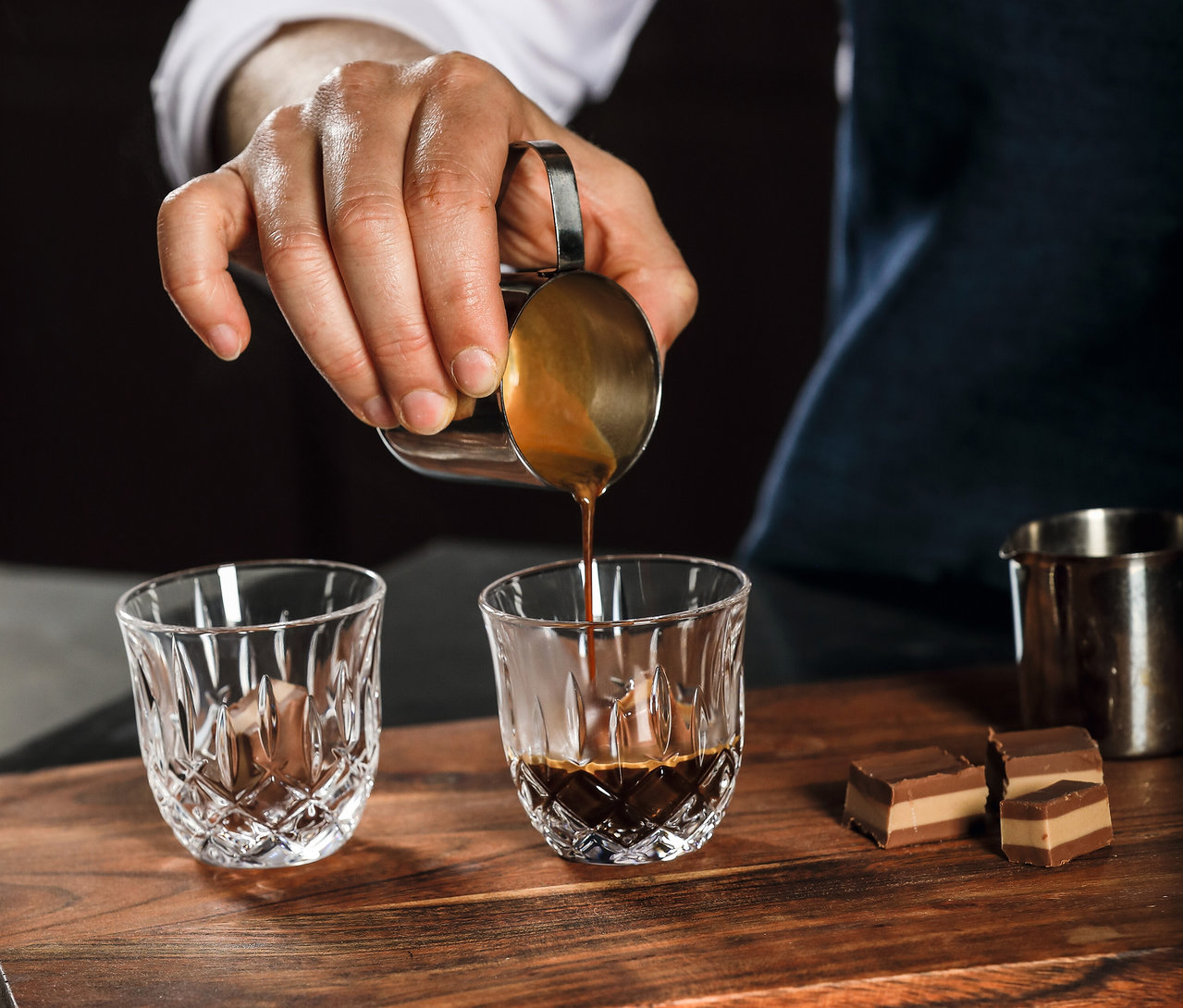 Mann gießt Espresso in ein Nachtmann Espressoglas »Noblesse« auf einem Holztisch. Pralinen liegen daneben.