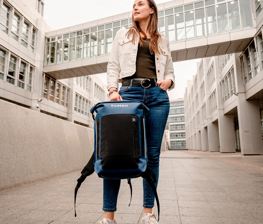 Frau hält blauen Tambu Rolltop-Rucksack RANA vor einem Gebäude.