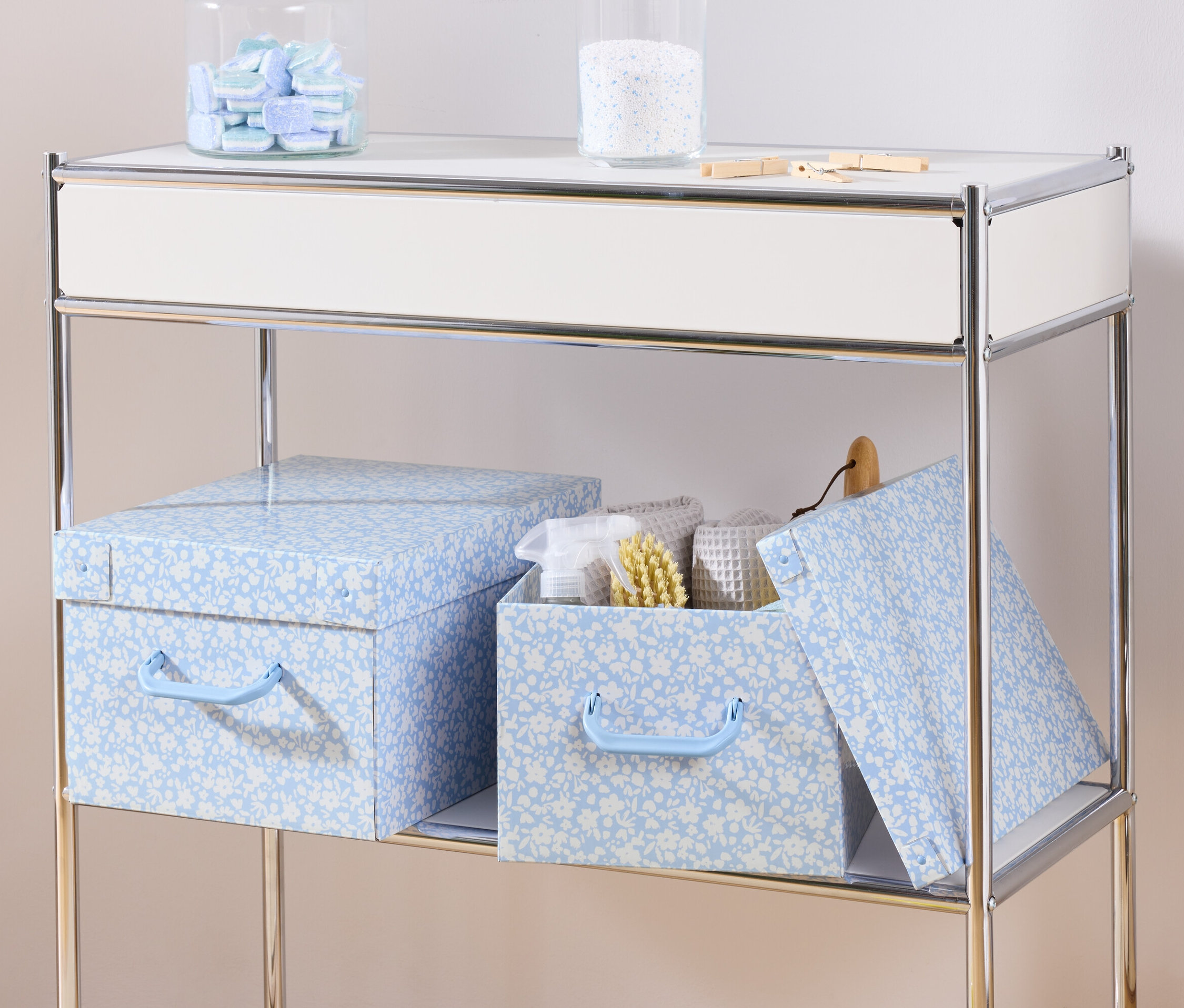 Auf einem Regal stehen zwei blaue Aufbewahrungsboxen mit Deckel und eine Aufbewahrungsbox mit Deckel mit weißen Handtüchern und einer Bürste.