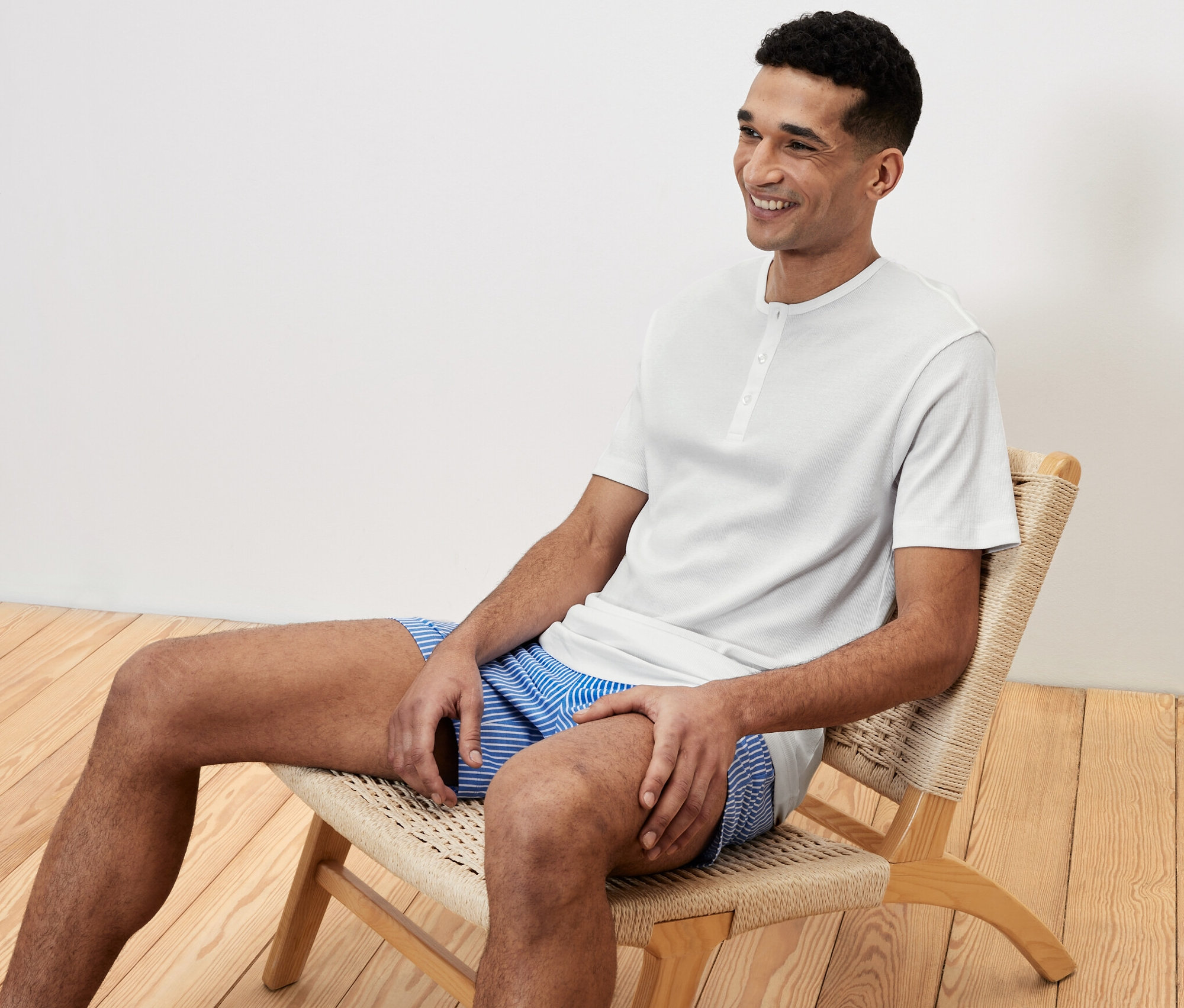 Mann sitzt in einem Shorty-Pyjama-Set auf einem Stuhl und lächelt.