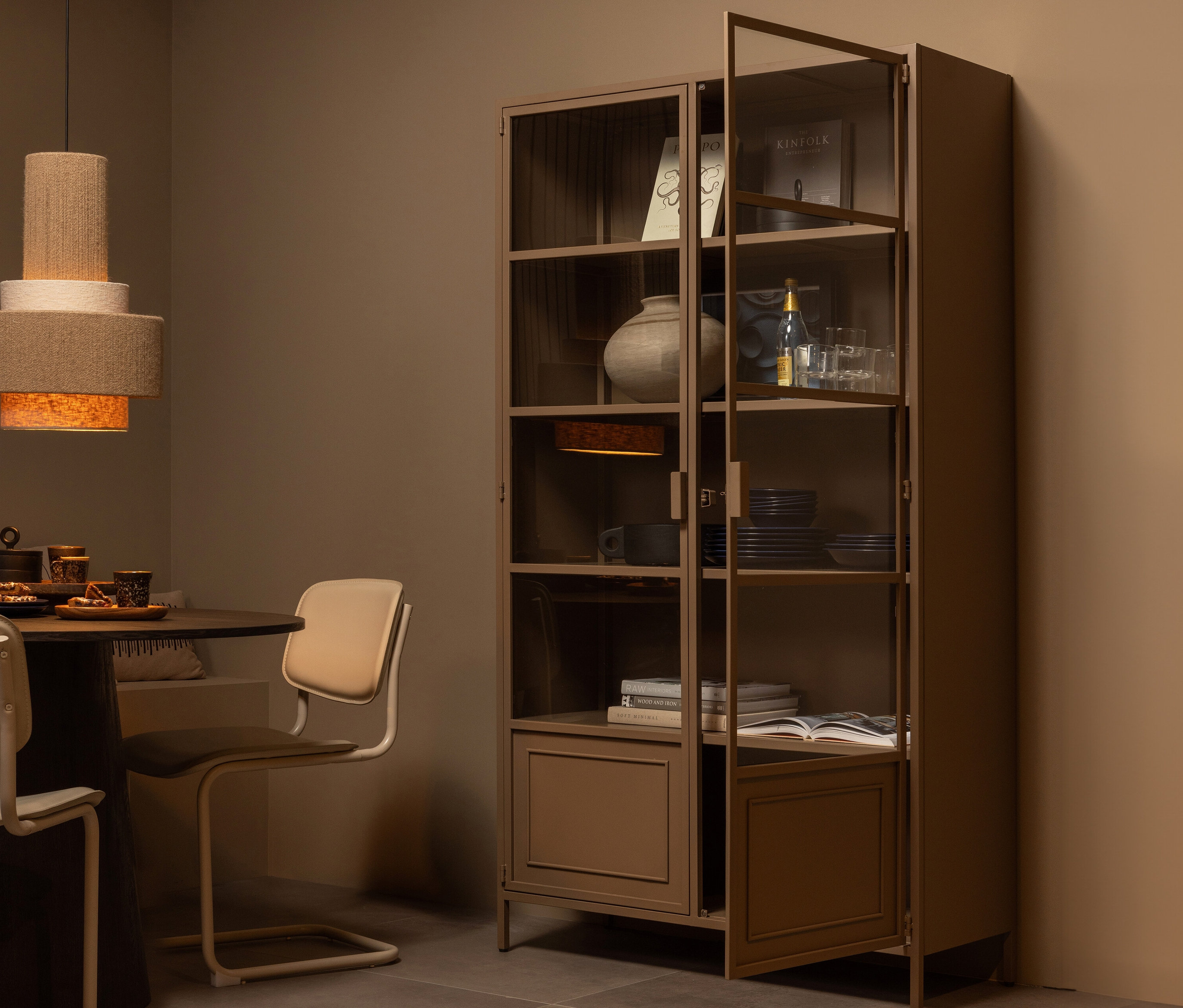 Eine teilweise geöffnete, bronzefarbene Woood »Ronja« Metallvitrine XL steht in einem Raum neben einem Esstisch.