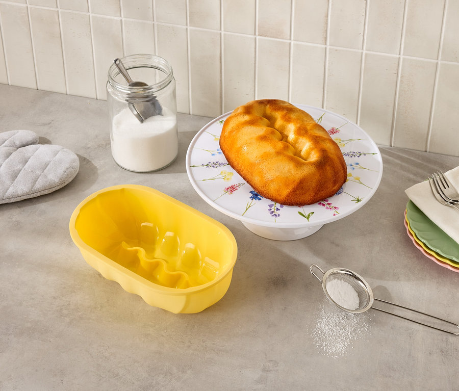 Auf einer Küchentheke stehen ein Gugelhupf auf einem Blumenständer, eine gelbe Gugelhupfform, ein Glas Zucker, ein Sieb und Topflappen.