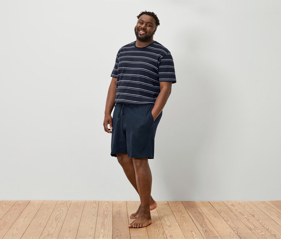 Ein lächelnder Mann steht barfuß vor einer weißen Wand und trägt ein gestreiftes T-Shirt und marineblaue Shorts.