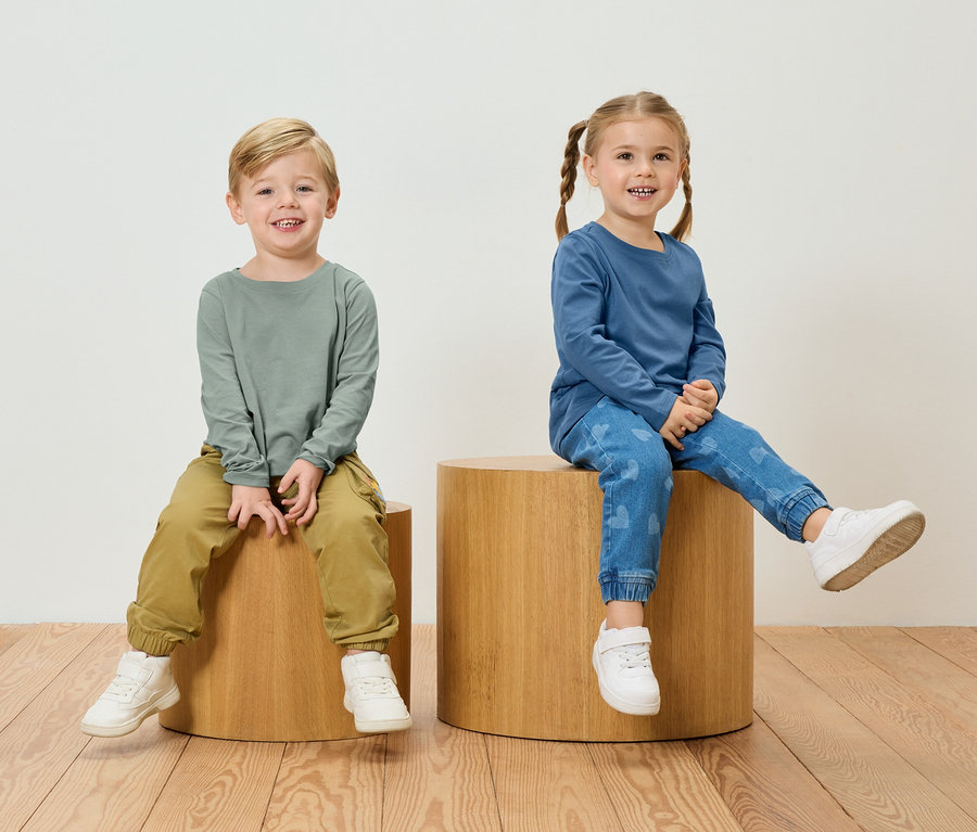 Ein Junge in einem grünen Langarmshirt und Khakihosen sitzt neben einem Mädchen in einem blauen Langarmshirt und Jeans mit Herzmuster auf Holzhockern.