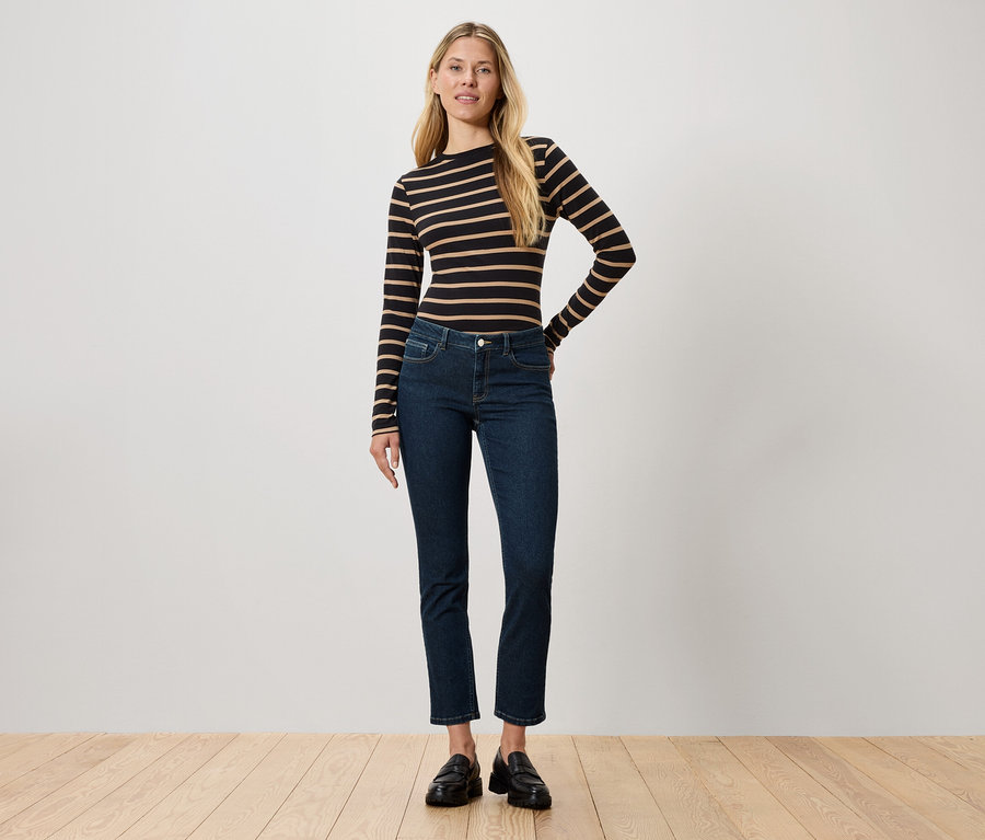 Eine Frau steht und trägt ein langärmliges, schwarz-braun gestreiftes Oberteil, dunkle Jeans und schwarze Schuhe.