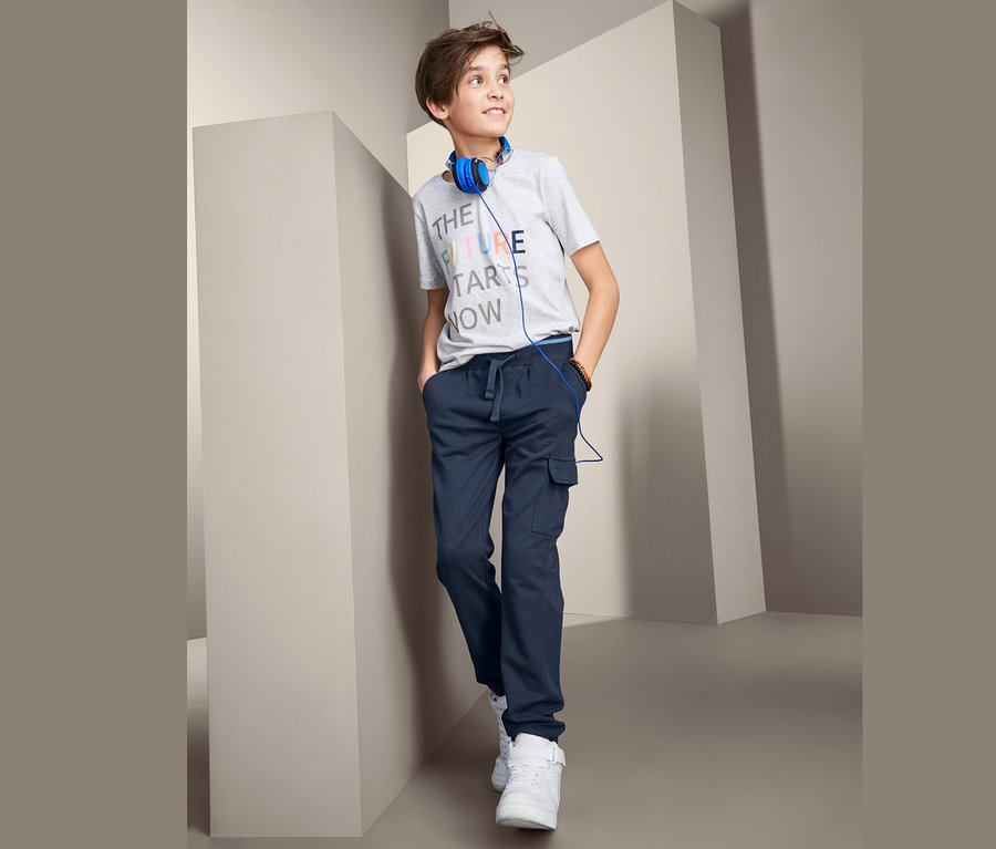 Ein Junge in einem grauen T-Shirt und einer dunkelblauen Schlupfhose mit Taschen, trägt blaue Kopfhörer und weiße Schuhe.