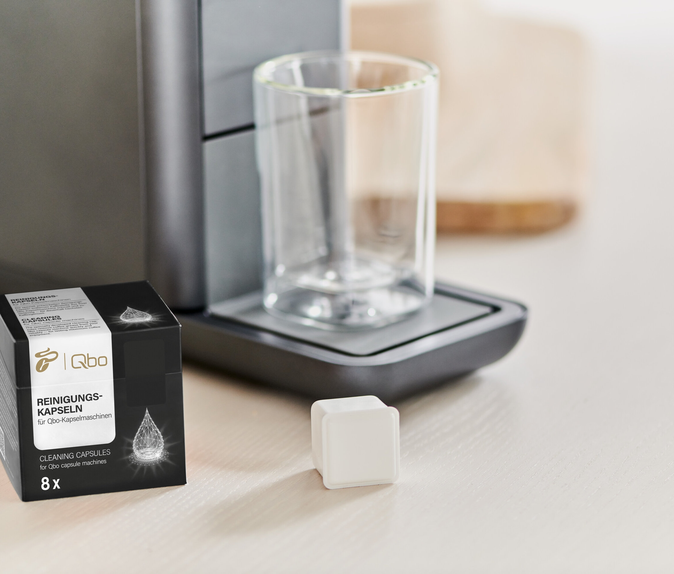 Eine Packung Qbo Reinigungskapseln steht neben einer Kaffeemaschine und einem Glas.