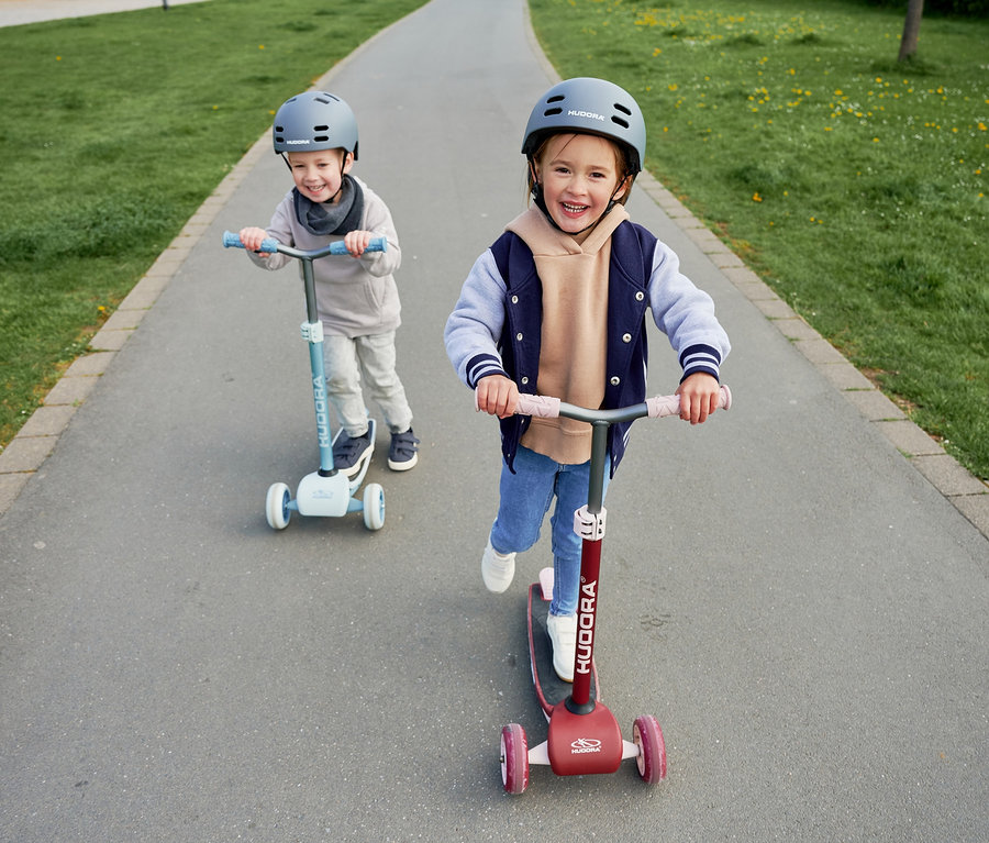 Zwei Kinder fahren auf dem Gehweg mit HUDORA Tri-Scootern. Ein Scooter ist bordeauxrot, der andere blau.
