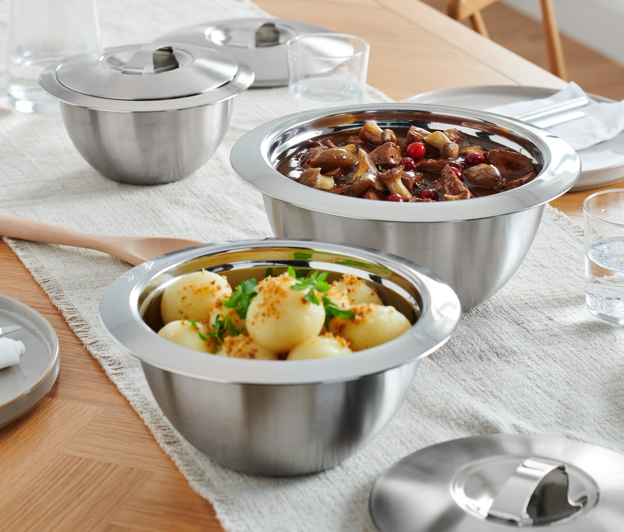 Auf einem Holztisch stehen drei Servier-Thermoschüsseln mit Edelstahldeckel, Teller und Wassergläser.