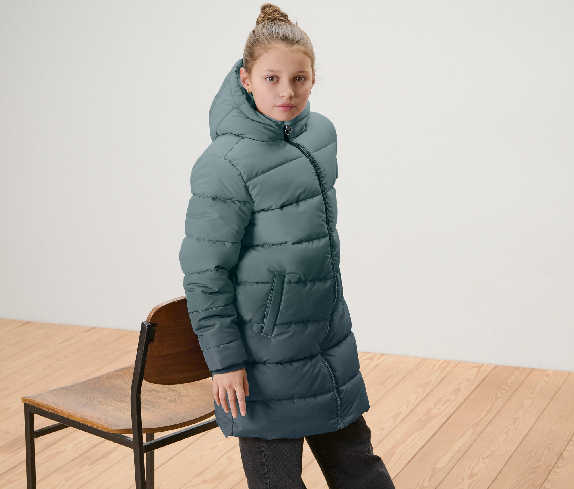 Mädchen trägt Kinder-Thermomantel mit Farbverlauf und steht neben einem Holzstuhl.