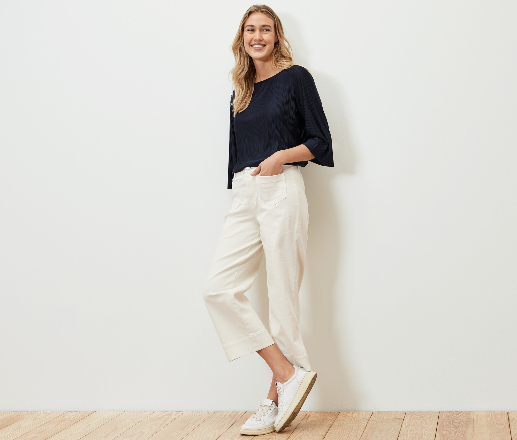 Frau posiert in Plissee-Blusenshirt, weißen Hosen und Turnschuhen.